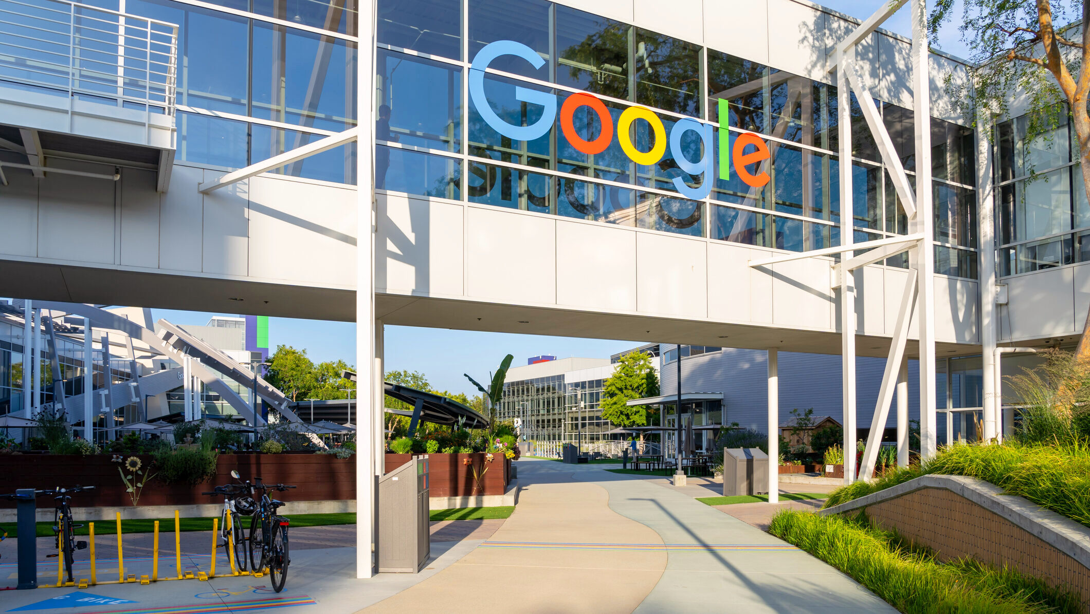 An elevated pedway with the Google logo is shown above a sidewalk path at Alphabet's headquarters in Mountain View, California.