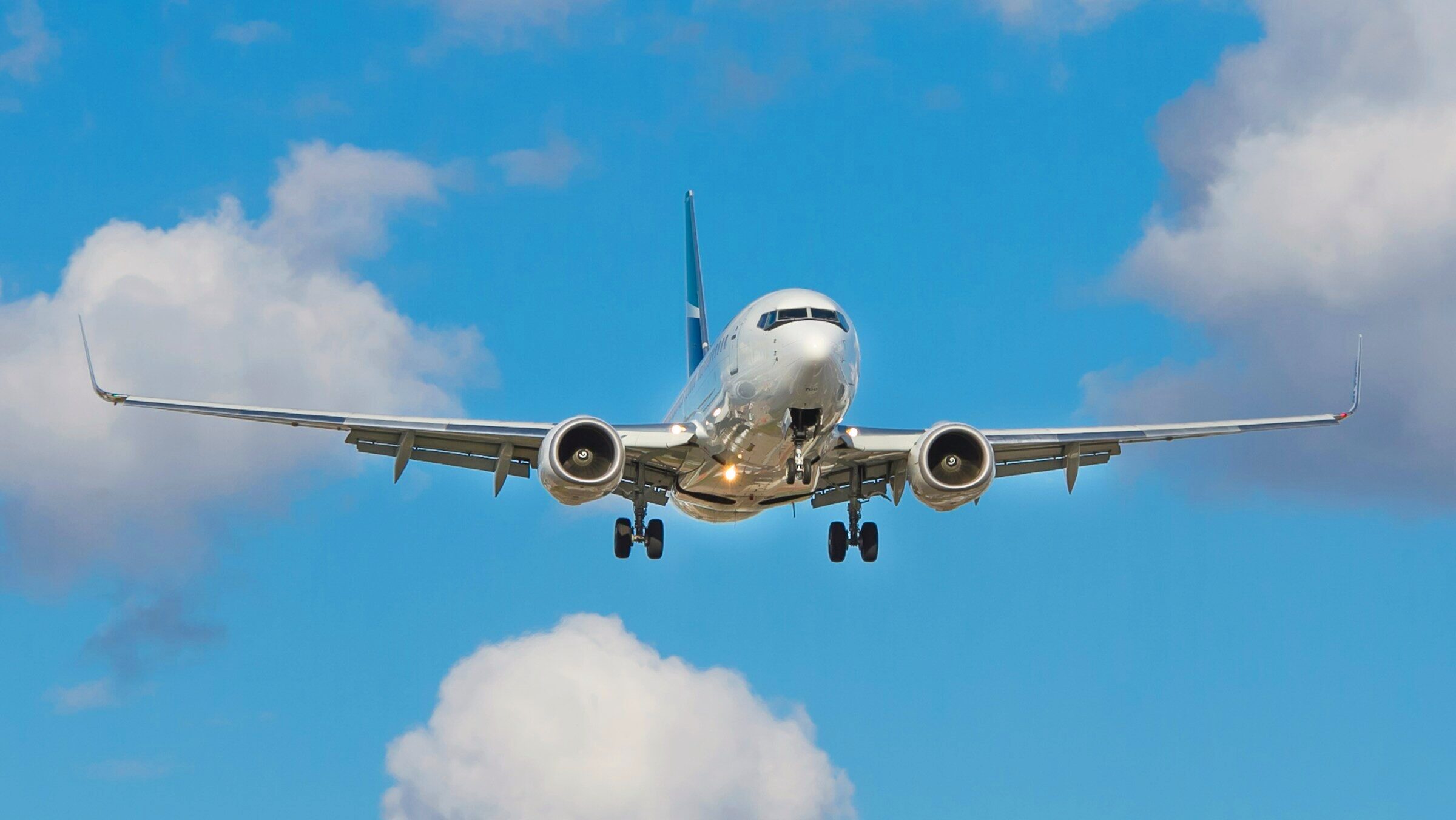 A flying Boeing commercial jet with its landing gear deployed descends in front of a blue sky with white clouds.