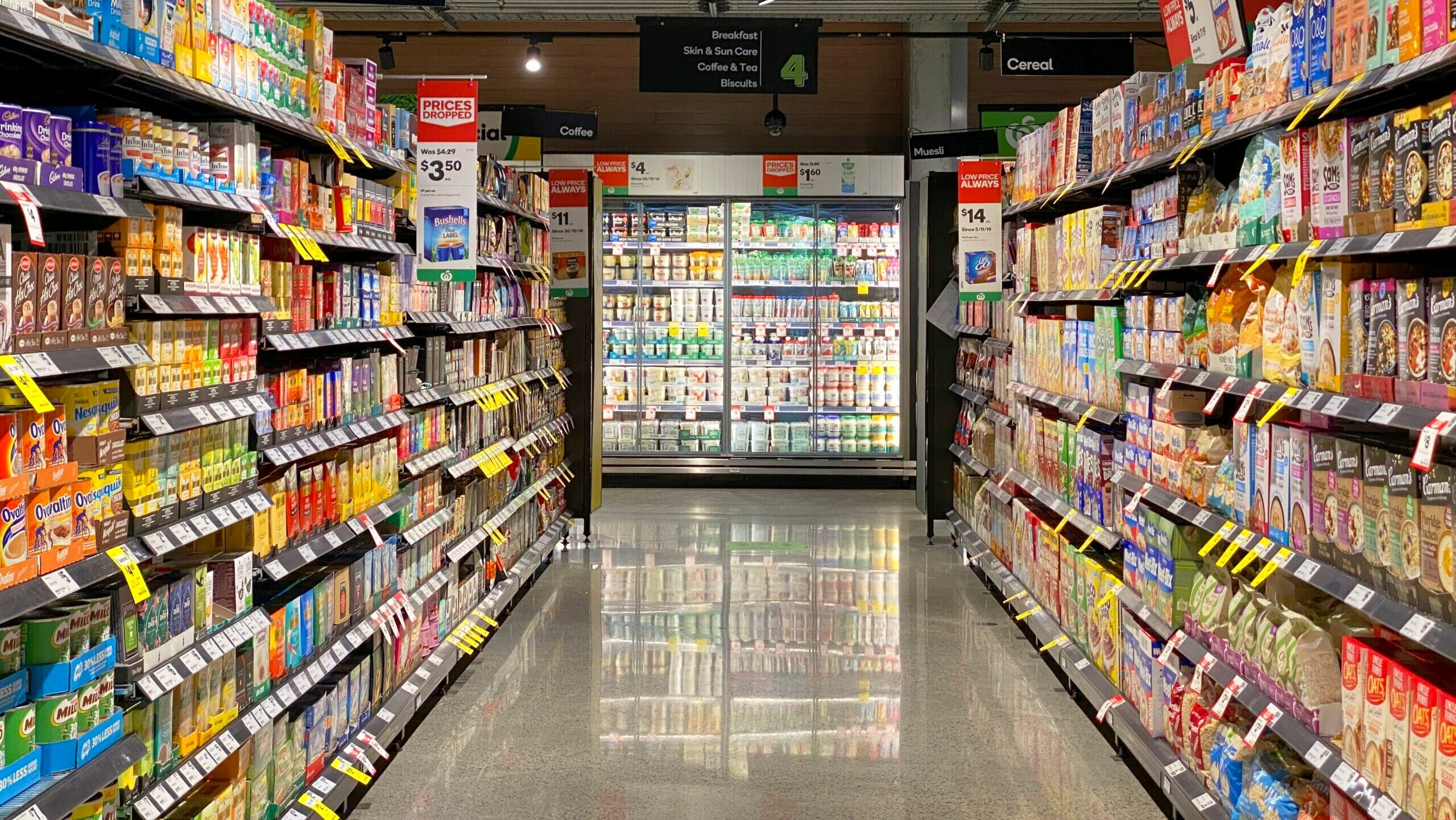 Two grocery store shelves containing boxed food products are shown on either side, leading to a refrigerator that contains chilled food products.