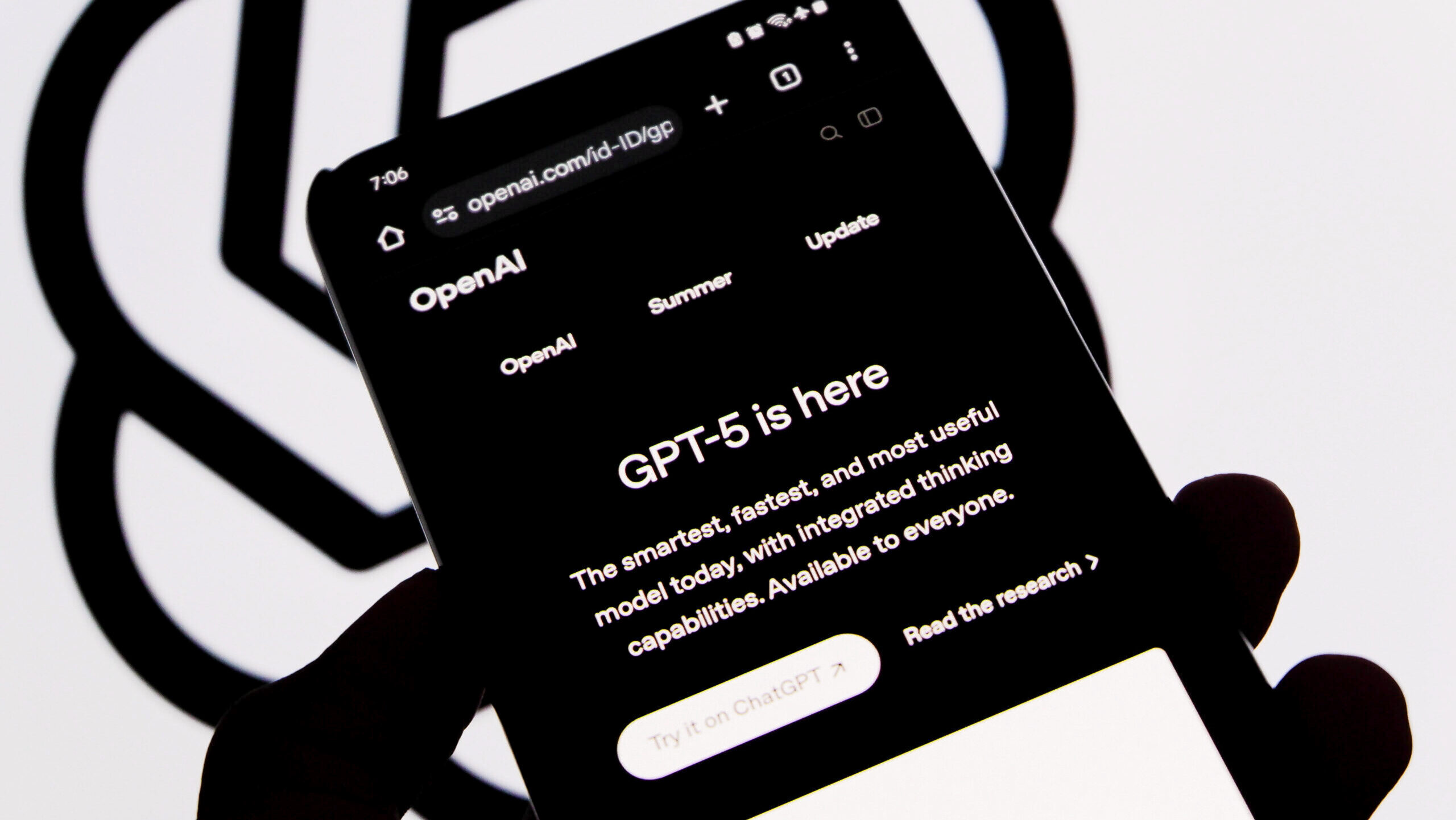 In this photo illustration, a person holds a smartphone showing the Introducing GPT-5 interface in the ChatGPT app in front of a blurred OpenAI logo background.