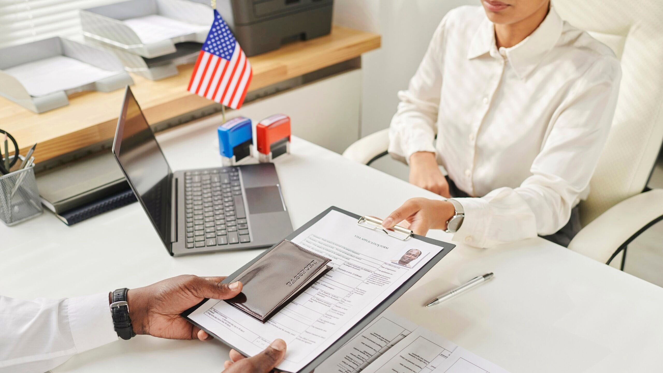 A person hands an immigration application for to a worker at a desk with an American flag on it.