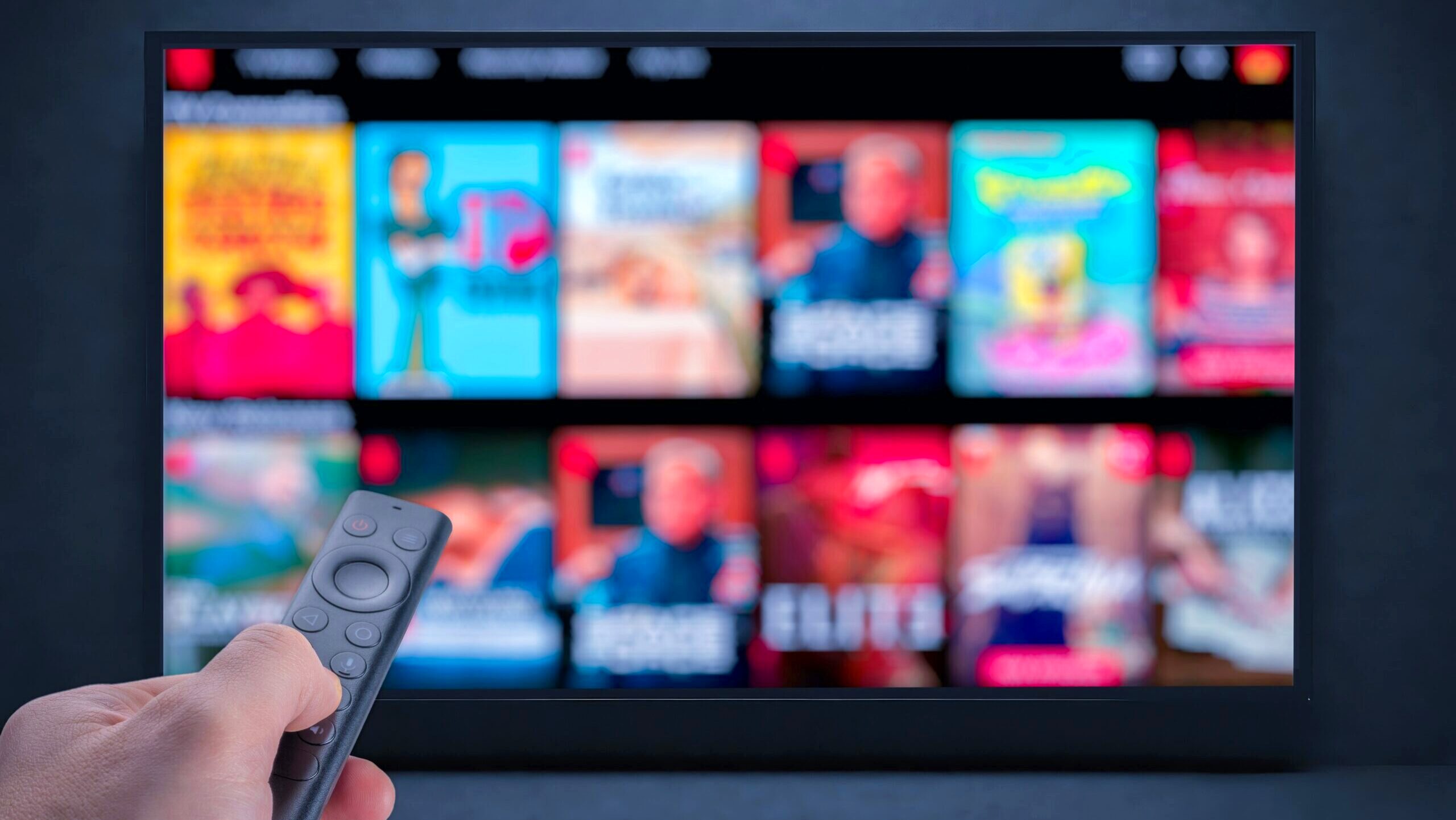 A person's hand is shown holding a remote control in front of a screen in the distance on which a Netflix menu is displayed.