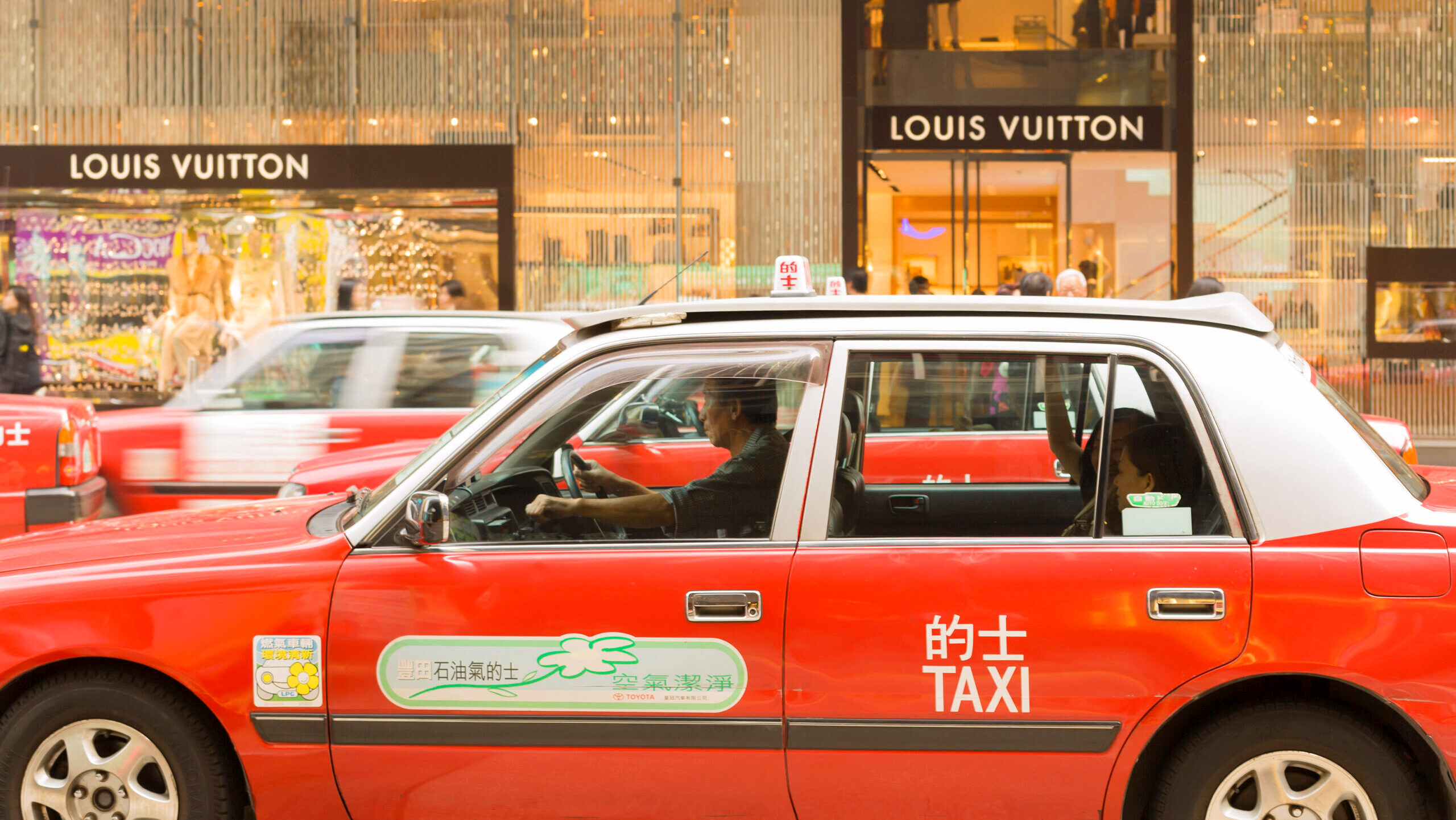 A traditional red taxi drives by a Louis Vuitton store in Hong Kong, China.