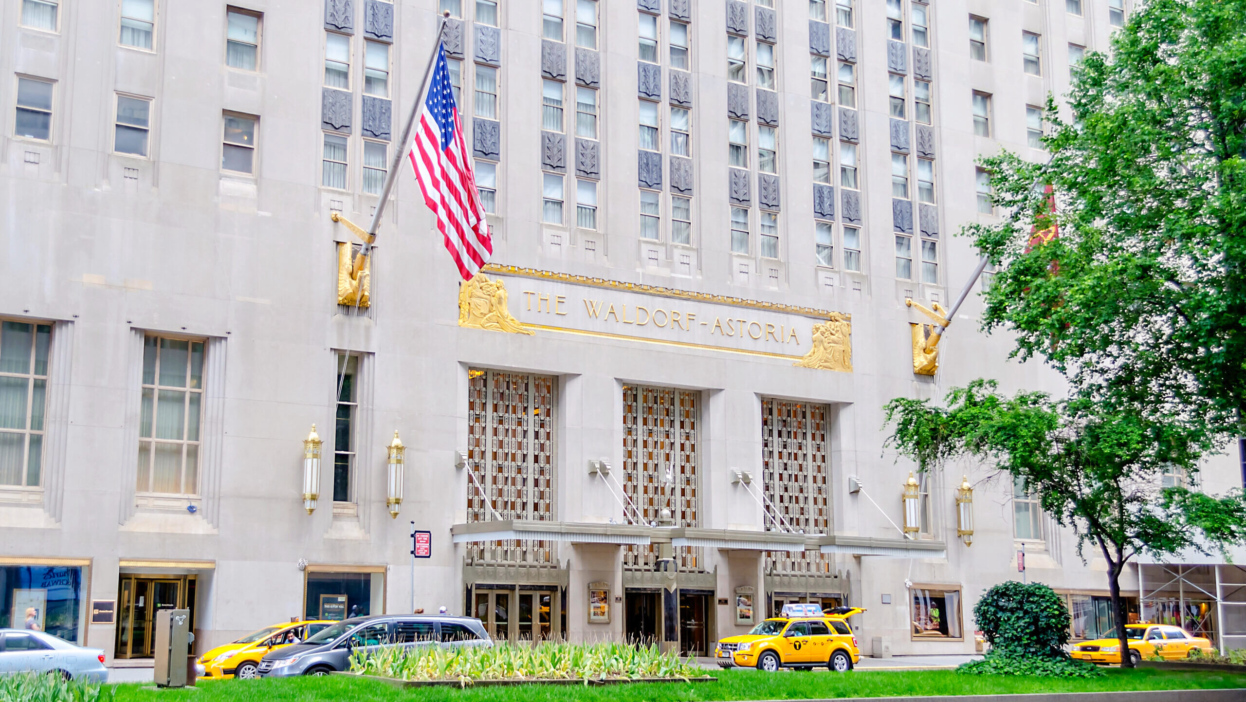 The exterior of the Waldorf Astoria Hotel in New York City is shown with cars and taxis passing by out front.