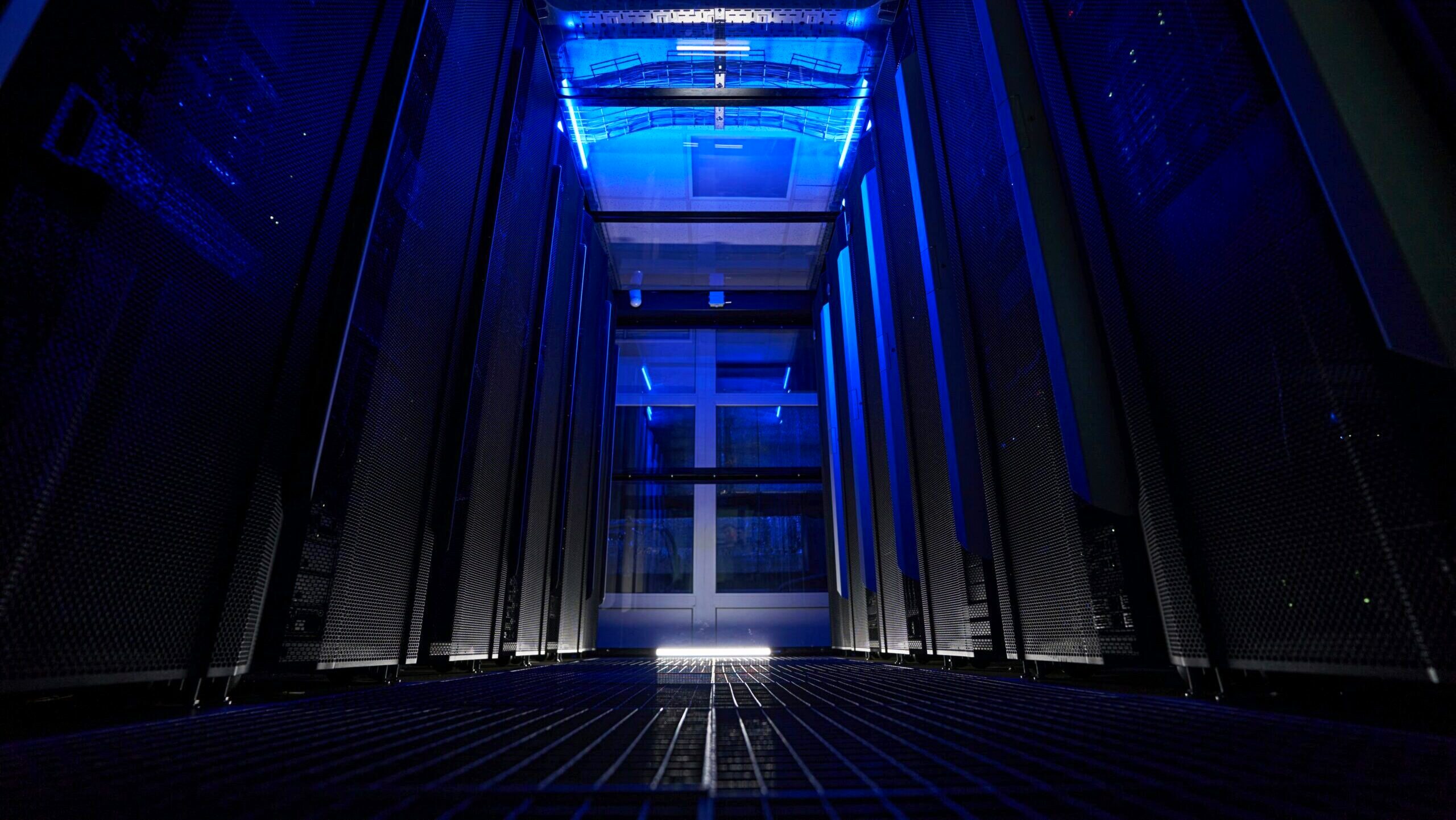A lowly lit server room inside a data center is shown with racks of data storage hardware in metal cabinets.