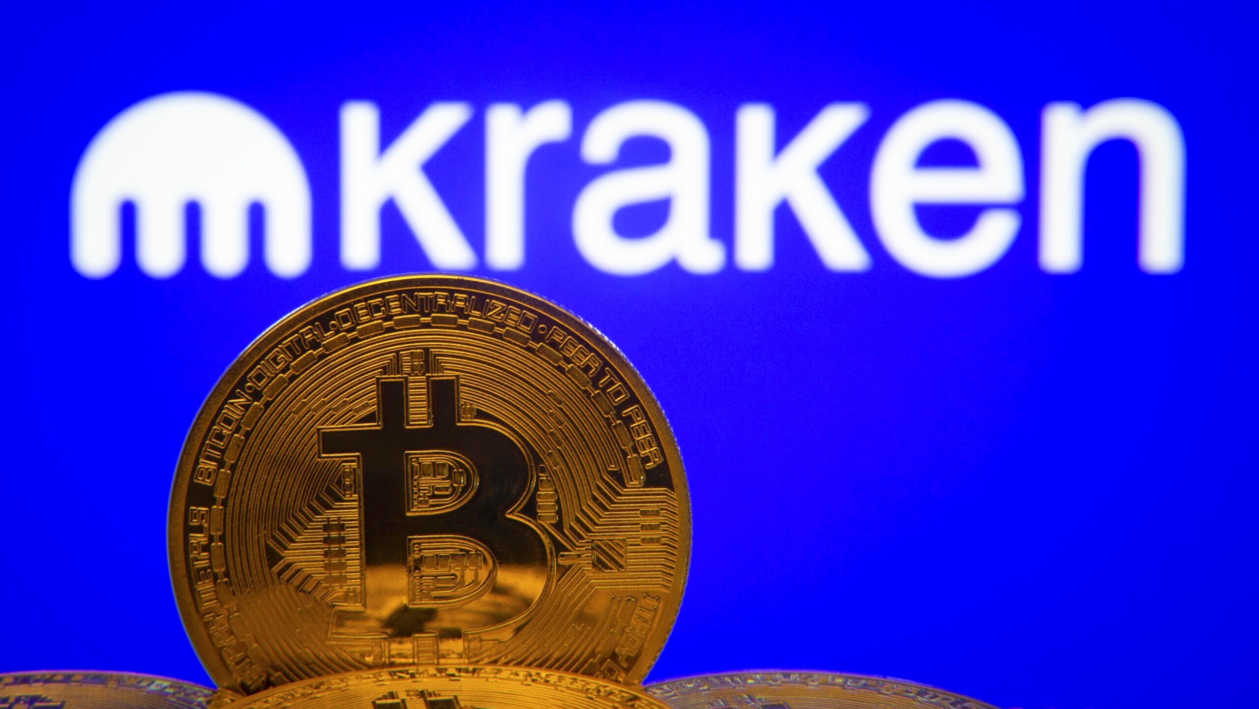 A golden representation of a bitcoin stands in the foreground before the Kraken logo.