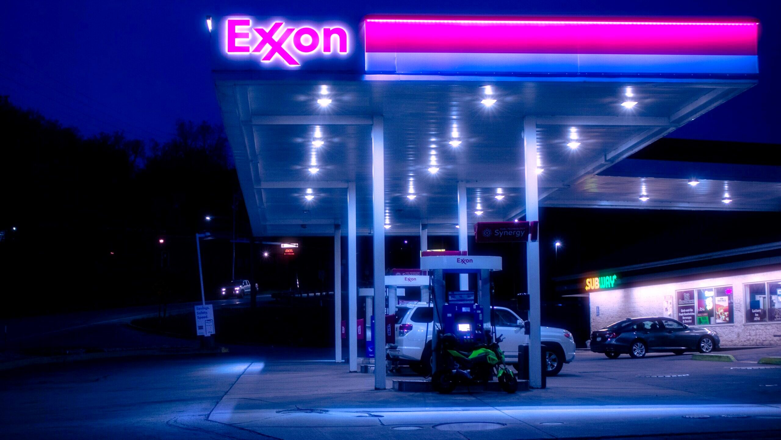 An Exxon-branded gas station with several vehicles parked is shown at night.