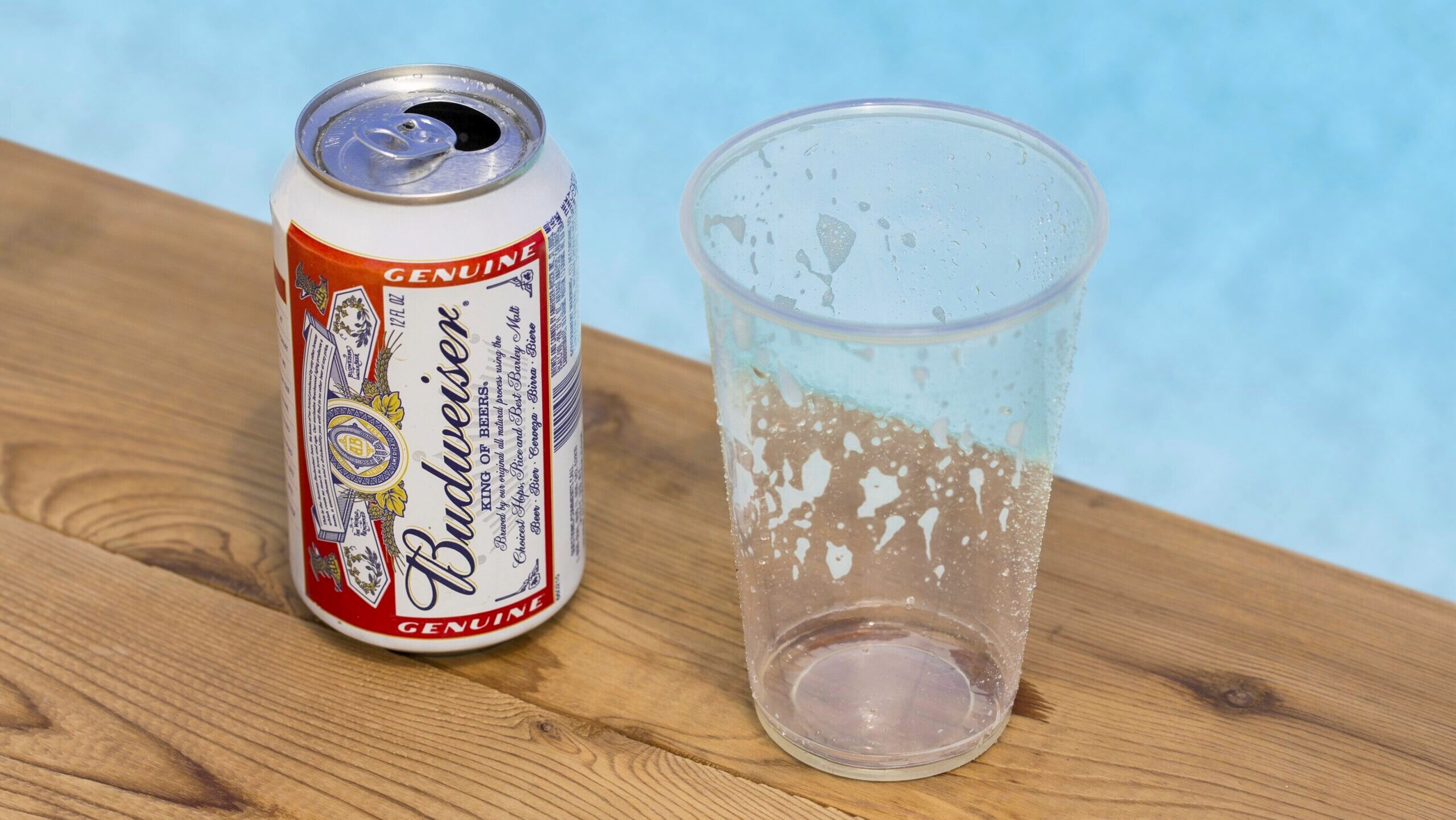 A can of Budweiser beer is shown next to a plastic disposable cup on a wooden table by a swimming pool.