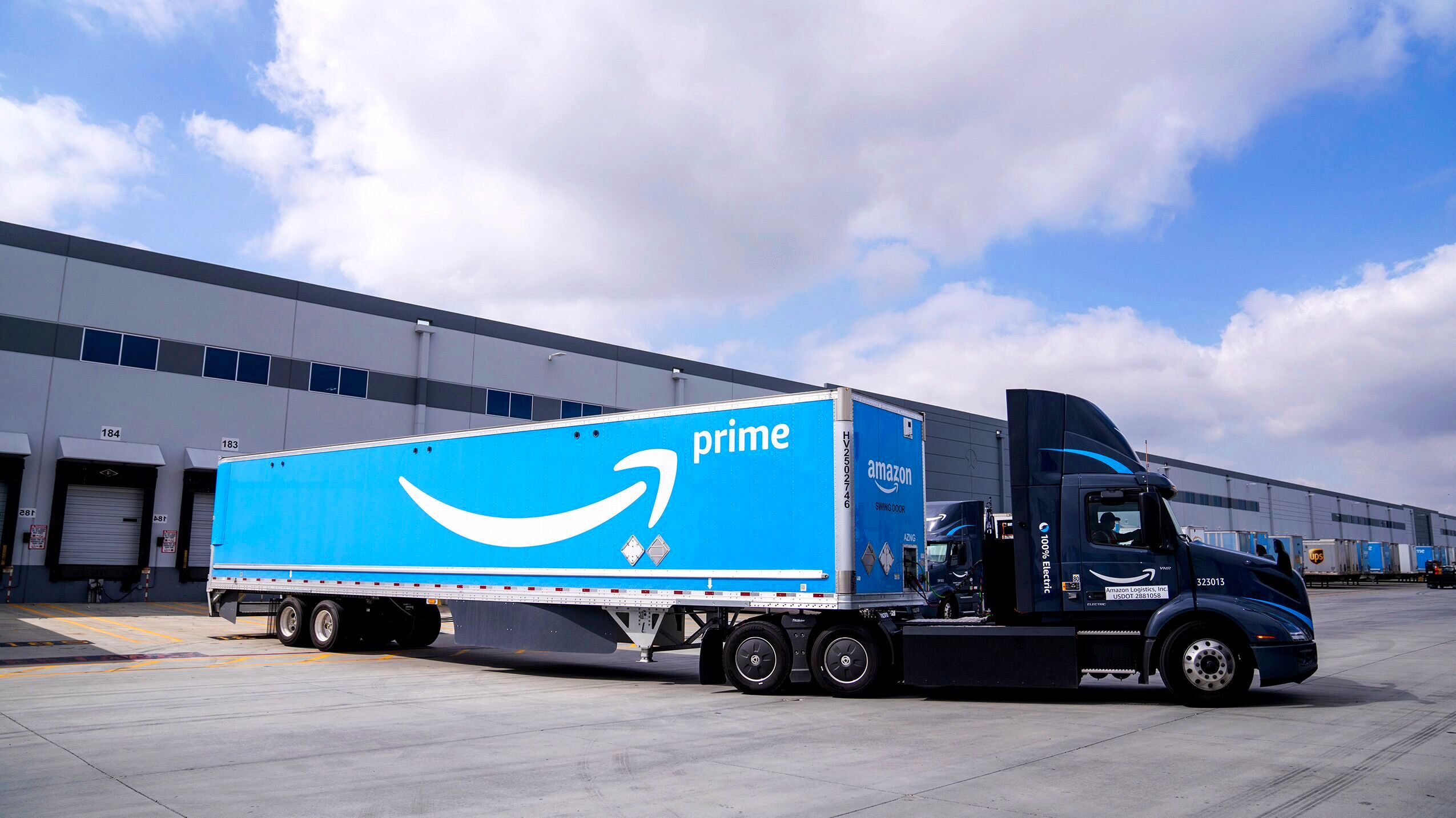 An Amazon freight truck is shown driving outside of a fulfillment warehouse.