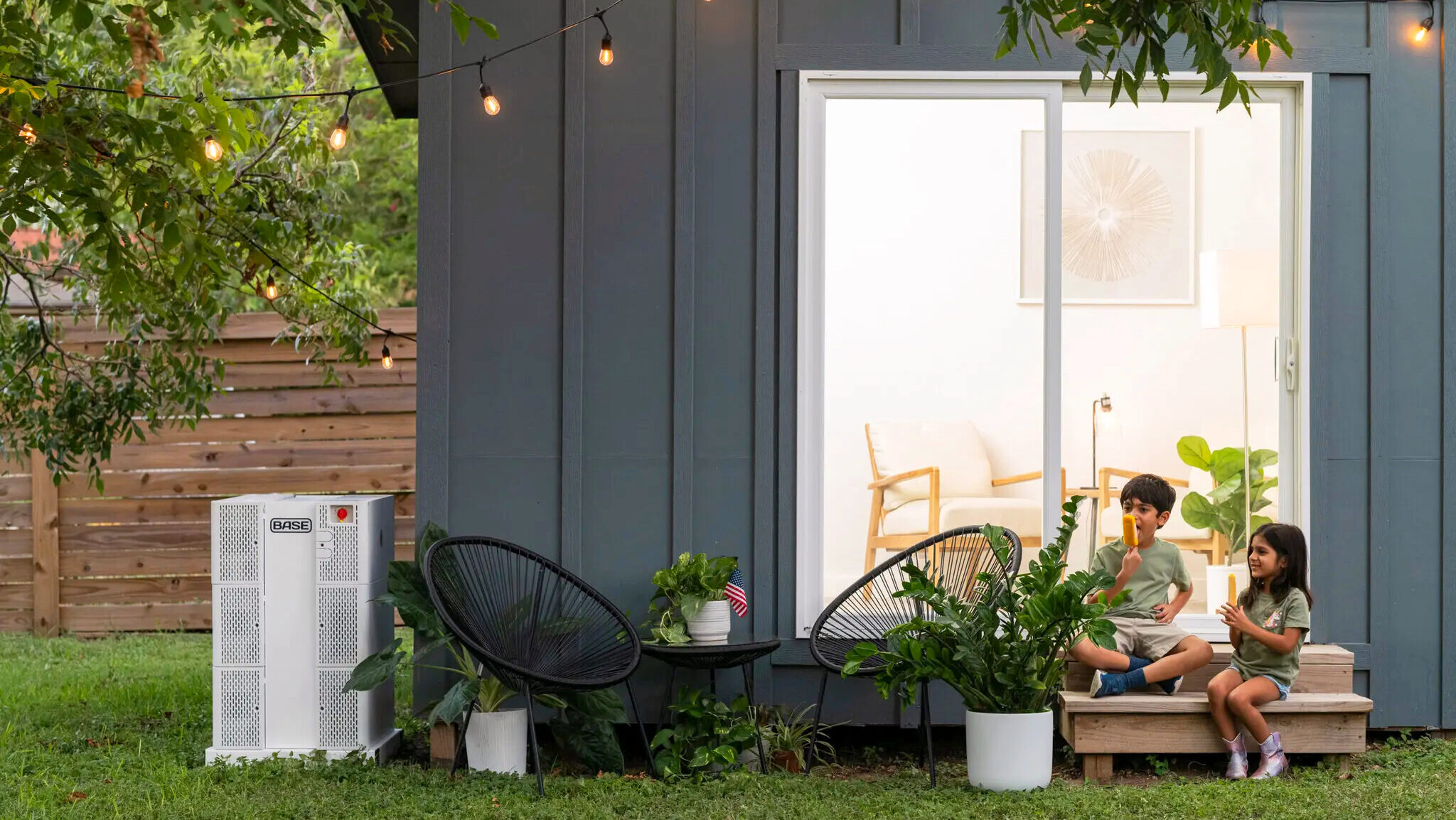 A Base Power home battery is shown next a residence where two children are enjoying frozen popsicles on the doorstep.
